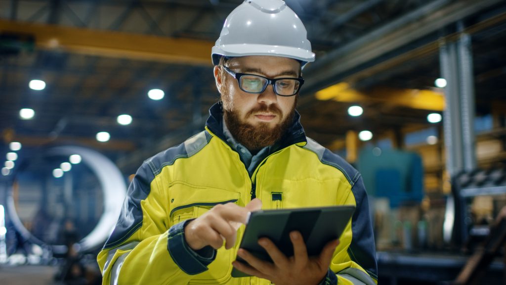 Man wearing a helmet and safety equipment while using a tablet in a manufacturing plant