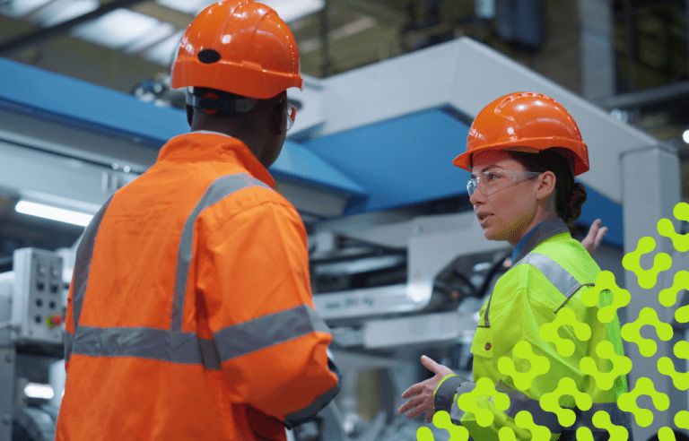 Two factory works in safety gear having a conversation while standing in a manufacturing facility.