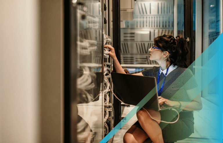 woman IT professional working on cables inside a server rack while kneeling down with a laptop on her knee.
