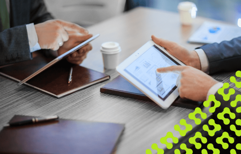Close up of two men discussing data on two tablets, with notebooks and plastic coffee cups around them.