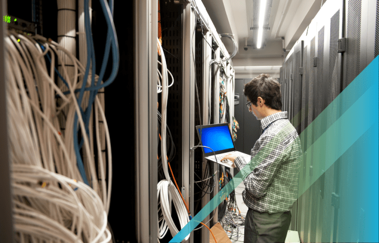 IT professional typing on a laptop in a server room surrounded by cables.