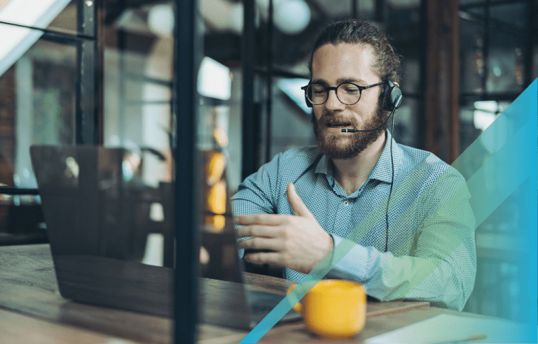 Man in eyeglasses and a button-up shirt working on a laptop wearing a headset and a yellow mug sitting beside him.