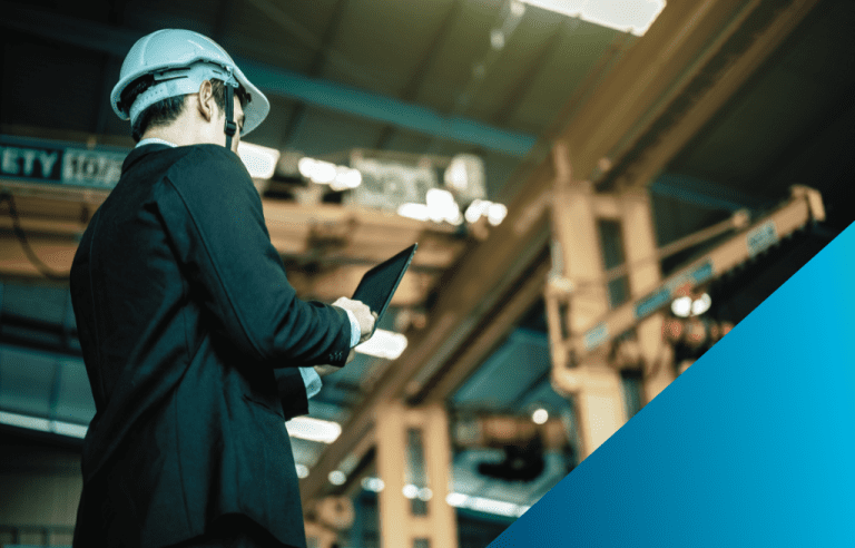 A man in a suit and a hard hat is viewed from behind, holding a tablet while inspecting an industrial facility with large overhead cranes.