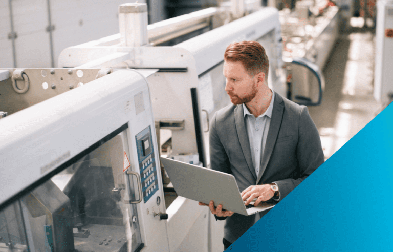 A man in a business suit holding a laptop while interacting with a piece of automated manufacturing equipment on a factory floor.