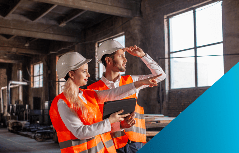 Two supervisors in hard hats and orange safety vests inspect an industrial site, with the woman holding a tablet and pointing.