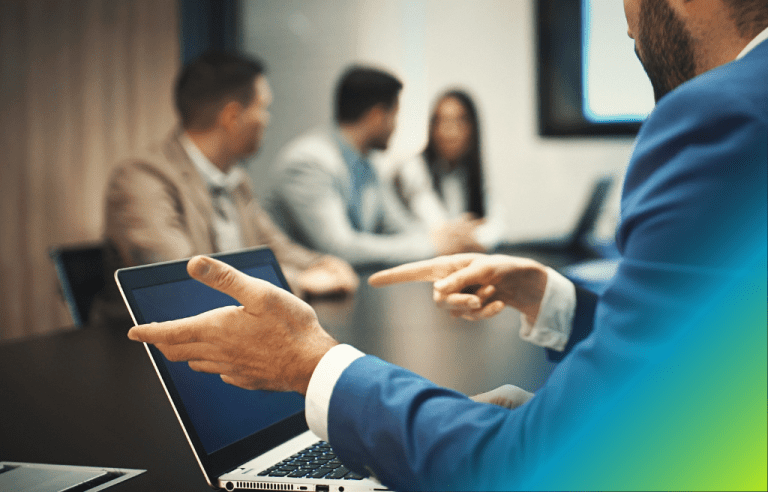 Close up of a man's hands pointing at a laptop screen while a business meeting takes place in the background.