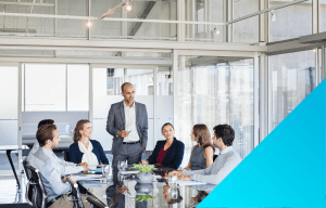 conference room full of people in business attire, one guy in the middle is standing holding a pen.