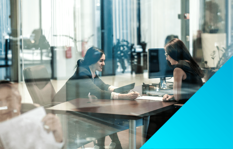 Shot through a glass window, two business women discussing information on a piece of paper.
