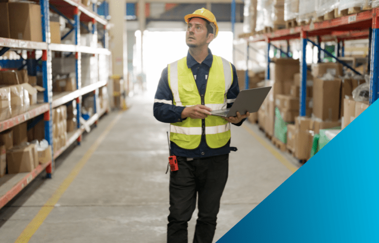 A man wearing a yellow hard hat and reflective vest holding a laptop while looking slightly upwards in an inventory warehouse.