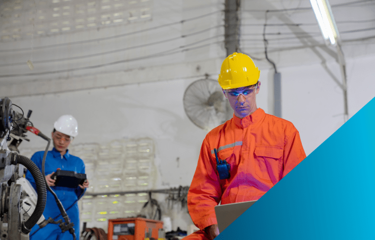 A male worker in an orange jumpsuit and yellow hard hat uses a laptop, while a female colleague in a blue jumpsuit and white hard hat uses a tablet in the background of a factory floor.
