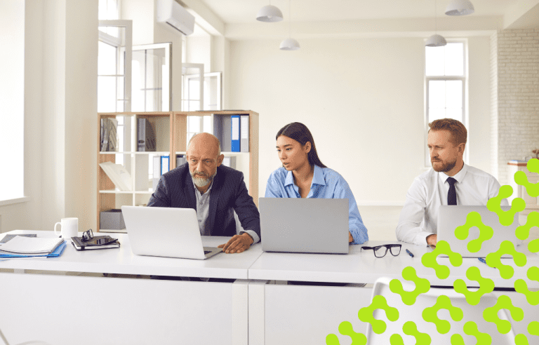 Three office professionals, two men and a woman, sit at a workstation using laptops to collaborate.