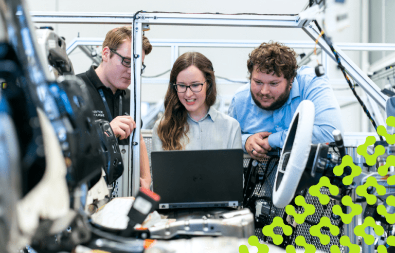 Three engineers are gathered around a laptop on the factory floor, working on a partially assembled product.