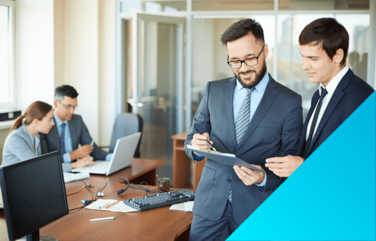 two pairs of business employees, one looking at a laptop and the other discussing documents on a clipboard.