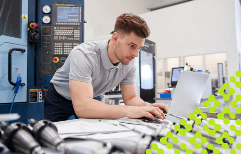 Man typing on a laptop in a room full of technical controls and parts.