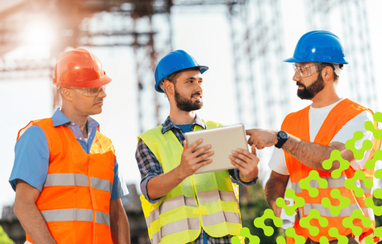 Three construction workers in hard hats and safety vests are gathered, with one pointing at a tablet while speaking to the other two.