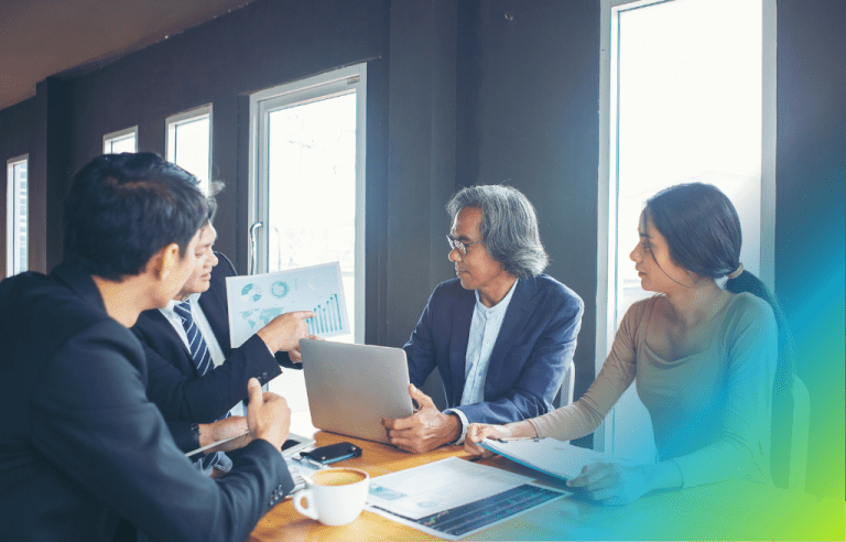 4 people discussing at a business meeting, one man holding up a piece of paper displaying statistics, while another man holds his laptop.