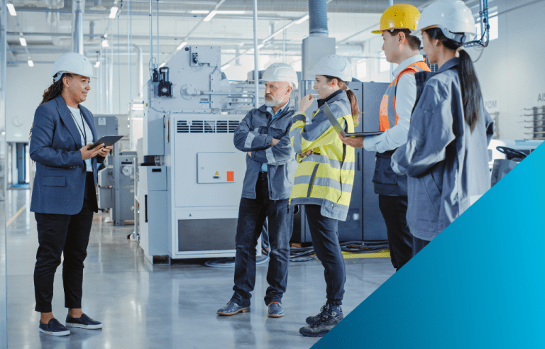 A diverse team of manufacturing professionals, wearing hard hats and safety gear, conduct a meeting on the factory floor in front of an industrial machine, using a tablet for digital data review.