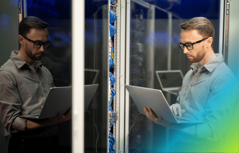 A man with glasses using a laptop in a server room, reflected in the glass of a server rack.