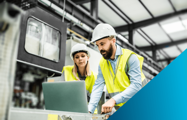 Two factory workers in hard hats and vests review data on a laptop near an automated machine.